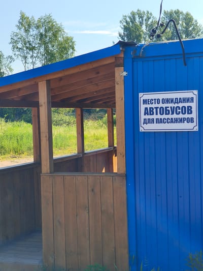 The image shows an outdoor bus stop shelter. The shelter has a wooden roof and posts, with a wooden floor and sides. A blue corrugated metal wall of a building forms the right side of the shelter. On this blue wall, there is a rectangular sign with a white background and blue text. The sign reads: "МЕСТО ОЖИДАНИЯ АВТОБУСОВ ДЛЯ ПАССАЖИРОВ", which translates to "BUS WAITING AREA FOR PASSENGERS". The bus stop appears to be located in a rural or semi-rural area, with green vegetation and trees visible in the background. The sky is clear and blue. The location is identified as Kady, Russia.