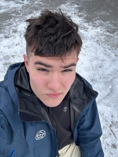 A young adult male is depicted in a high-angle self-portrait taken outdoors in Anger, Austria. The background consists of a partially snow and ice-covered ground.

The subject has short, dark brown, textured hair with shaved sides and a fringe. His skin is light-toned with minor facial redness. He possesses dark brown eyes and maintains a neutral, direct gaze toward the camera.

He wears a dark blue outer jacket, which features a white circular logo depicting a stylized animal on the left chest. A black fleece garment is visible beneath the outer jacket, displaying a white Timberland tree logo on its left chest. A portion of light-colored trousers is visible at the bottom of the frame. No other individuals or significant objects are present in the immediate vicinity.
