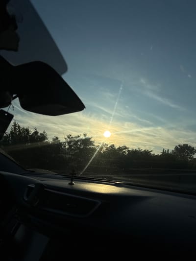 The image captures a view from inside a car, looking through the windshield towards a sunset or sunrise. The sky is a gradient of blue and yellow, with wispy clouds. The sun, a bright white orb, is positioned towards the left side of the frame, casting a warm glow and lens flares. In the distance, a dark silhouette of trees forms a horizon line. The interior of the car is visible in the foreground, with the dashboard and a rearview mirror. A small crucifix figurine is placed on the dashboard. The image appears to have been taken on a road in or near the city of Trușeni, Moldova.