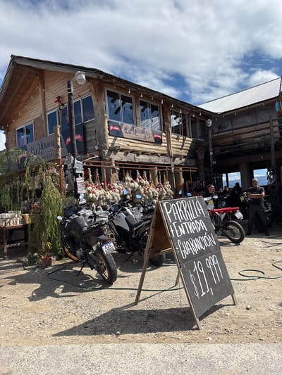 The image shows a two-story, wooden building in Villa La Merced, Argentina, likely a restaurant or shop, with a sign that reads "La Ponderosa Fiambreria" in Spanish. The building has multiple windows on the upper level and a corrugated metal roof. Outside the building, rows of what appear to be strings of onions or garlic are hung. Several motorcycles are parked in front, obscuring the view of the building. A chalkboard sign stands near the motorcycles, displaying handwritten text in Spanish, including the words "ENTRADA," "PORRU," and possibly a price of $19,999. Two people are visible on the second-story balcony. The ground is gravel. The sky is partly cloudy.