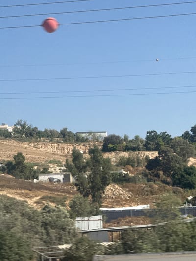 This media file captures a bright, sunny day in a hilly, vegetated area, likely in al-Qasoum Regional Council, Israel. The image is oriented vertically, with the sky taking up a significant portion of the left side. Several power lines are visible, crisscrossing the blue sky.
In the sky, two spherical objects are present. One is a small, light-colored sphere, appearing somewhat distant. Closer and more prominently featured on the left side is a larger, reddish-pink sphere, which is slightly blurred, suggesting movement or a shallow depth of field. This could be a balloon or a similar lightweight object.
The right side of the image depicts a steep, rocky hillside covered in sparse vegetation and trees. Buildings and structures are partially visible nestled into the hillside or on its upper edges. Some appear to be modern, with metal siding, while others are less defined. The terrain is dry and dusty, indicative of a warm climate. The presence of trees suggests it is not a desert landscape, but rather a semi-arid or Mediterranean environment.
The overall scene appears to be a view from a moving vehicle or elevated position looking across a landscape dotted with human infrastructure. There are no visible people, and the activity implied is incidental, such as the presence of the spheres in the sky. The bright light suggests it is daytime, likely late morning or afternoon, with clear weather. No text is visible in the image.