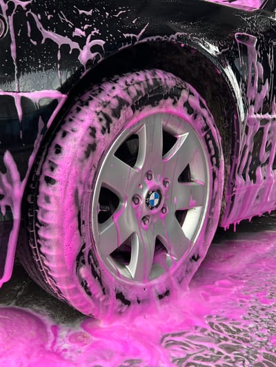 This image depicts a detailed close-up of a dark-colored automobile undergoing a wash process in Velyki Hai, Ukraine. No individuals are visible in the frame.

The vehicle's rear wheel arch, lower quarter panel, and a significant portion of the wheel and tire are covered in vibrant pink, viscous soap foam. The foam adheres to the vehicle's dark, reflective surface, forming numerous streaks and rivulets as it drips downwards. The multi-spoke alloy wheel, painted silver, prominently displays a blue and white BMW logo on its center cap. Visible details include lug nuts and the tire's tread pattern, which also retains significant foam.

Beneath the vehicle, the ground—a dark, wet paved surface—is covered in a pooling and spreading layer of the same pink foam and water. The lighting suggests an outdoor environment during daylight hours.