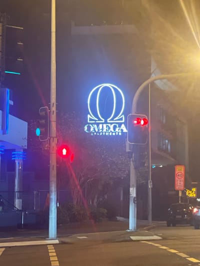 The image depicts a nighttime street scene at an intersection in Brisbane, Australia, specifically near the Omega Apartments. A large, illuminated neon sign for "OMEGA APARTMENTS" is prominently displayed on a building, featuring a stylized Omega symbol in blue and white. A traffic light is visible on the right side of the frame, showing red lights. To the left, another traffic light displays a green light. A speed limit sign for a "SCHOOL ZONE" with "40" and specific hours is also visible. Cars are present on the road, and streetlights and building lights illuminate the surroundings, casting reflections on the wet asphalt. The environment appears urban with some vegetation, possibly trees, visible behind the apartment building.