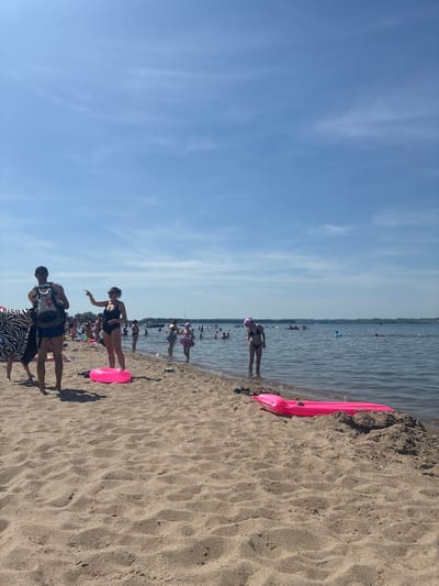 This is a vertically oriented, eye-level shot of a sandy beach with people enjoying the water on a sunny day. The sky is a clear blue with wispy white clouds. The water is calm and stretches out to the horizon, where a distant shoreline is visible. Several people are wading in the shallow water or swimming further out.
On the beach, a bright pink inflatable pool float is partially submerged in the sand near the water's edge. In the foreground, three individuals are standing on the sand. One person wears a black swimsuit and a pink hat, pointing towards the water. Another person, carrying a backpack, is dressed in a black and white zebra-print sarong and a t-shirt. The third person is mostly obscured.
Further back, more people are visible on the beach and in the water, some with floats. The scene appears to be a popular recreational spot, likely during daylight hours given the bright sunlight and clear sky. The mood is relaxed and leisurely, suggesting a typical summer day at the beach. The location is identified as Rewa, Poland, which is a coastal town. There is no visible text in the image.