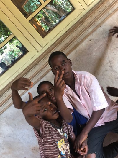 Three dark-skinned children pose on the floor near a window. The child at the top wears a pink shirt and dark pants. The second child has on a blue shirt. The third child is in a striped shirt and holds a lollipop in their mouth. All three are making hand gestures, showing two fingers in front of their eyes. Part of a wooden-framed window is visible on the left, reflecting trees and foliage. A partial hand and foot are visible in the top of the frame.