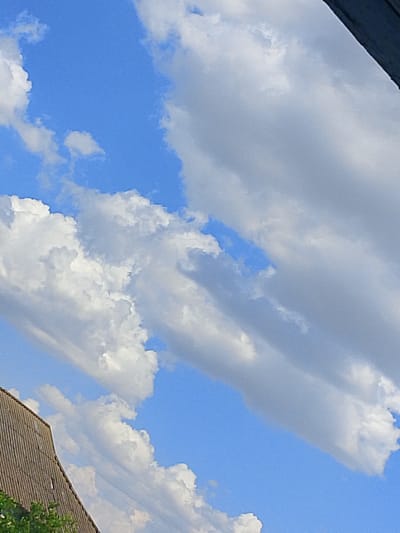 This is a low-angle, eye-level shot of a bright blue sky with scattered, voluminous white and grey clouds. The clouds are dynamic, with some appearing fluffy and others streaky. A portion of a grey, corrugated roof of a building is visible in the lower left corner, with some green foliage at the very bottom edge. The image appears to be taken outdoors in Dobra Krynytsia, Ukraine.