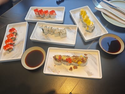 The image shows a table with several plates of sushi and a small bowl of soy sauce. There are at least three different types of sushi rolls visible: one topped with red roe, one sprinkled with black and white sesame seeds, and one topped with what appears to be mango and a creamy sauce. The sushi is arranged on rectangular white plates with a decorative pattern. In the background, a fork and knife are resting on a napkin on a larger plate. The scene appears to be indoors, likely in a restaurant, given the presentation of the food and cutlery. The lighting suggests it could be daytime, though no specific time can be determined. No people are clearly visible in the frame, only a hint of a person's arm at the top left. The overall impression is a meal being served or enjoyed. There is no visible text in the image.