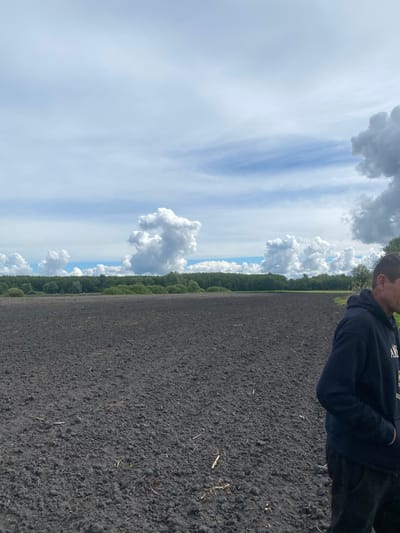 The image shows a landscape in Pidluky, Ukraine. A person stands on dark, tilled soil. A line of green trees separates the field from a sky filled with white and gray clouds. The sky transitions from cloudy to blue as it ascends in the image. The person is wearing a dark hoodie and pants. The view is slightly angled and the subject is at the right of the frame.