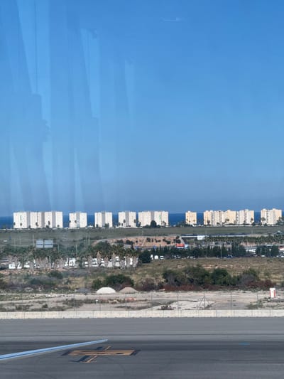 The image depicts an aerial view taken from what appears to be an airplane window, showing a section of the city of Elche/Elx, Spain. The scene is divided into three prominent sections: the sky/sea, a line of buildings with intervening vegetation, and an airport runway. The sky/sea occupies the left side of the image and is a bright blue. A series of white or light-colored buildings, likely apartments or hotels, lines the coast. Between these buildings and the airport runway is a strip of vegetated land with trees and smaller buildings. The airport runway takes up the right side of the image and is gray, with markings indicating it is an active airport space.