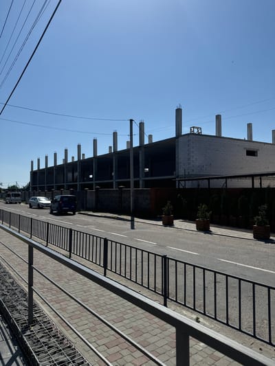 The image shows a construction site with an unfinished building under a clear blue sky. The building has a concrete frame and appears to be several stories high, with exposed rebar at the top of some pillars. Several cars are parked on the asphalt road next to the construction site, including a dark blue van and a white car. The road has white lane markings. To the right of the road, there is a paved walkway with stone tiles and a metal fence. Planters with small evergreen trees are lined up along the building. In the distance, there are power lines. The overall scene suggests a sunny day in a semi-urban or industrial area, possibly in Sarny, Ukraine, given the provided location context. There are no visible people or text in the image.