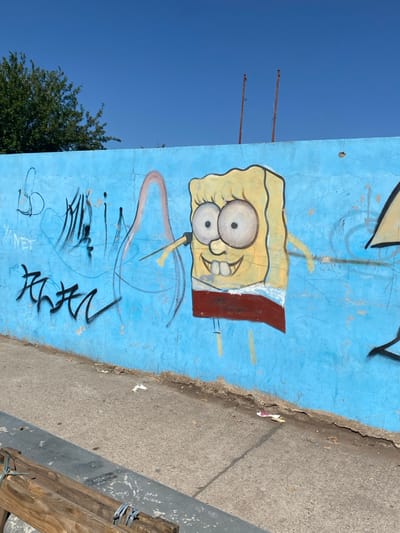 A cartoon image of SpongeBob SquarePants adorns a light blue, cracked wall covered in additional graffiti. The character sports a wide-eyed, buck-toothed grin and is drawn with thin lines. Below SpongeBob, more abstract graffiti is scribbled in black. The wall is adjacent to a grey sidewalk with visible debris. A portion of a wooden structure is visible on the right side of the sidewalk. A clear blue sky and the top of a tree frame the left side of the image.