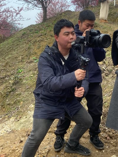 Two men operate a camera rig outdoors, likely for filming. One man holds the camera and stabilizer, wearing a dark jacket and gray jeans, while the other lies behind him, supporting the setup. Both wear dark shoes. The setting appears to be a natural environment with flowering trees, suggesting a rural or park location.