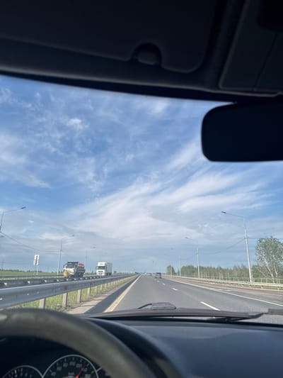 The image displays a daytime view from the interior of a vehicle, looking forward onto a multi-lane highway. Elements of the car's interior frame the scene, with the roof lining and sun visor visible in the top-left and a partial view of the dashboard and steering wheel on the right.

The highway stretches into the distance under a bright blue sky, which features scattered, wispy white clouds, indicating clear and fair weather. The road has distinct white lane markings and is bordered on the right by a metal guardrail. Beyond the guardrail, a green verge leads to a dense line of trees, suggesting a natural or rural environment.

Street lights or utility poles line the side of the highway, receding into the background. Several large trucks are visible in the distance, traveling on the road. The scene captures a moment of highway travel, consistent with a location such as Усть-Сыны, Russia. No discernible text is visible within the image.