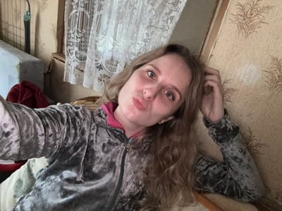 A young woman with blonde hair and blue eyes is taking a selfie in a room. She is wearing a grey velour hoodie with a pink and blue lining. Her left hand is touching her hair, and her lips are pursed. The background shows a window with a white lace curtain, a wall with floral wallpaper, and a portion of a mattress and a red cloth on the left side. The setting appears to be domestic, possibly in a residential building in Severnyy, Russia.