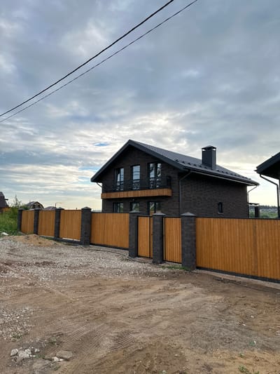 The image depicts a residential property in Bol'shaya Mos', Russia, under an overcast sky. A prominent two-story house constructed of dark brown or black brick stands centrally, featuring a dark pitched roof with a visible chimney and a black metal balcony railing on the upper floor. The roof is equipped with dark gutters and downspouts.

In the foreground, a newly installed wooden fence, light brown in color, encloses the property. The fence posts are constructed from dark brick matching the house. A large sliding gate and a smaller pedestrian gate, both made of vertical wooden slats, are integrated into the fence. The ground directly in front of the fence is unpaved, consisting of dirt and gravel with numerous tire tracks.

In the background, other residential structures with dark roofs are partially visible among green vegetation, suggesting a developing suburban or rural residential area. Two dark power lines extend diagonally across the upper left quadrant of the cloudy sky. No individuals are present in the scene.