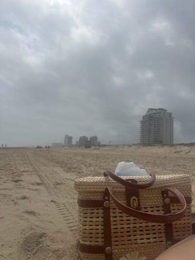 The image presents a low-angle perspective of a beach scene on South Padre Island, United States of America. In the foreground, a rectangular woven straw handbag with brown leather straps and metallic hardware is positioned on light-colored sand. A crumpled white plastic item is visible inside the bag's opening. A small section of human skin, indicative of a leg or foot, is present on the right edge of the frame. The sandy beach extends into the midground, displaying distinct tire tracks leading towards the horizon. In the background, a cluster of multi-story buildings, including a prominent white high-rise with numerous balconies on the right, defines the distant skyline of South Padre Island. Several small, indistinct human figures are visible on the far stretch of the beach. The sky is entirely covered with dense, gray overcast clouds.