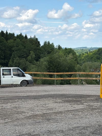 The image displays a white Ford Transit van parked on a gravel surface, with a wooden fence in the background. The fence has multiple horizontal planks with "TLC" printed on them. Beyond the fence, a dense forest of green trees stretches across the landscape under a partly cloudy blue sky. The van is positioned on the left side of the frame, facing to the right. The surrounding environment suggests an outdoor rural or industrial setting. There are no people visible in the image.
