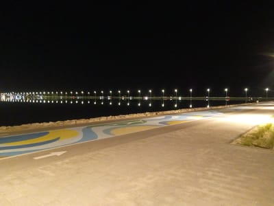 This image captures a nighttime scene along the waterfront in Al Khobar, Saudi Arabia, likely at Al Khobar Corniche or a similar coastal area. The foreground features a paved walkway with a decorative, wavy design in blue, yellow, and white. A white arrow indicating direction is visible on the walkway. Beyond the walkway, a low, rocky wall separates it from a body of calm water.
The water reflects a long, continuous line of streetlights that illuminate the distant shoreline. These lights appear as bright, distinct orbs, with their reflections creating a shimmering effect on the water's surface. The far background is dominated by darkness, with a few more distant lights suggesting the presence of buildings or a city skyline. The overall atmosphere is serene and modern, highlighting the well-lit urban landscape against the dark night sky. There are no identifiable people or specific objects in the immediate foreground or middle ground that are clearly discernible in detail.