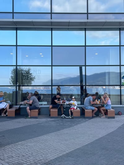 A group of people are dining at outdoor tables in front of a large glass-windowed building. The building has a modern design with a grid of large windows reflecting the sky and a distant body of water with mountains behind it. On the windows, text is visible, appearing to be "OUND OFFE". Several groups of people are seated at wooden tables with benches.
In the foreground, on the left, a woman wearing a straw hat and a white dress is seated at a table. To her right, a man in a grey t-shirt and dark shorts is eating. Further to the right, a man in a black t-shirt and shorts is also seated at a table.
In the middle of the scene, a young boy in a white t-shirt with a graphic print and red shorts is seated at a table with a scooter beside him. To his right, a couple is eating at a table. The man is wearing a grey t-shirt and blue shorts, and the woman is wearing a pink tank top and a denim skirt.
The ground in front of the tables is paved with interlocking grey bricks, arranged in a patterned fashion, with diagonal lines of darker and lighter grey. A shadowed area falls across the paving stones. The overall scene suggests a casual dining atmosphere in an urban or waterfront setting. The location is identified as N2 свх. Динской, Russia.
