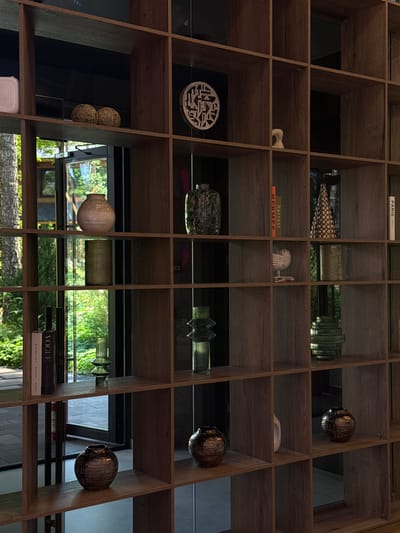 The image shows a close-up of a wooden bookshelf filled with various decorative objects. The bookshelf is constructed with numerous square compartments, creating a grid-like structure. The wood has a warm, brown, textured finish.
On the top shelf, to the left, is a white rectangular object. To its right are two woven spherical objects. Further to the right, on the same shelf, is a circular decorative piece with an intricate, geometric pattern.
The second shelf from the top contains a patterned vase with green and brown hues on the left. To the right of this vase, there is a white, abstract sculptural piece resembling a bird or swan.
The third shelf features a beige, textured vase on the left. To its right is a cylindrical, greyish vase. Further to the right, a tall, slender vase with a textured surface is visible.
The fourth shelf from the top has several items. On the left are two books, one with "VOGUE" written on its spine. Next to the books is a small, ribbed green glass object. Further to the right, there are two more decorative items, one resembling a small pine cone with metallic accents, and another cylindrical green vase.
The bottom shelf displays two dark, metallic vases with a ribbed texture, one on the left and another towards the right side of the shelf. In the center of this shelf, a white ceramic pitcher is visible.
The background through the compartments of the bookshelf reveals an outdoor environment with trees and greenery, suggesting the bookshelf is positioned near a window or opening to the outside. There are reflections in the dark backing of some of the shelf compartments, indicating a glazed or mirrored surface. The lighting appears to be natural, coming from the outdoor environment. The overall impression is of an interior space decorated with artistic and artisanal objects. The specific location is not identifiable from the visual information provided.