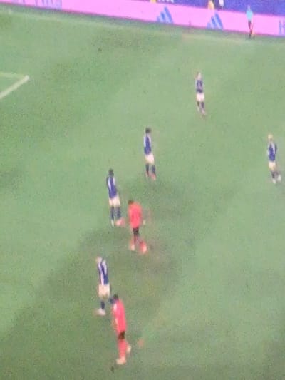 The image shows a blurred aerial view of a soccer game. Several players are visible on a green field with white markings. Some players are wearing blue and white uniforms, while others are wearing red uniforms. The players appear to be spread out across the field, with some in close proximity to each other, suggesting active play. The background shows a pink and purple advertising board or banner along the edge of the field. The image quality is low, making it difficult to discern specific details about the players or the location. However, based on the provided context, the scene is set in the city of Шипуново, Russia.