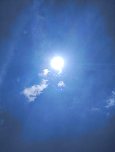 The image depicts an outdoor sky scene in Karai ward, Kenya. A brilliant, circular white sun, producing significant glare and a visible lens flare artifact to its lower right, is centrally positioned in the upper portion of the frame. The surrounding sky is a deep blue. Several small, detached white cumulus clouds are present, primarily in the lower-left quadrant, with additional darker grey cloud formations partially visible along the periphery of the frame, particularly in the upper-left and lower-right corners. No human figures, man-made structures, or ground-level objects are visible.