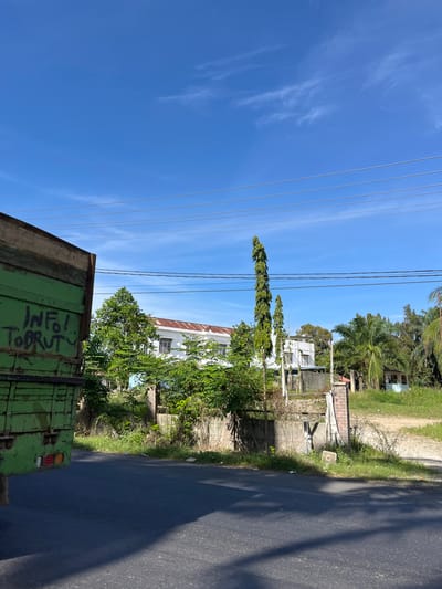 The image depicts a roadside scene in Bukitrata, Indonesia, under a clear blue sky with wispy clouds. A green truck, partially visible on the left, has graffiti on its side that reads "INFO! TOBRUTY". In the background, a white building with a reddish roof stands behind a low concrete wall and lush greenery. Several tall, slender trees and palm trees dot the landscape. A dirt path leads to a small, light blue structure further in the distance. Power lines are strung across the sky. The foreground shows a dark asphalt road with a shadow cast across it. The scene appears to be a sunny day in a semi-urban or rural setting.