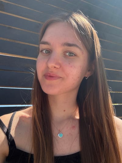A young woman with long brown hair and fair skin poses for a selfie. She has green eyes and light pink lips. She is wearing a silver necklace with a turquoise heart-shaped pendant and small earrings. The woman has some acne on her face. She is standing outside, with a black metal fence behind her. The location is unknown.