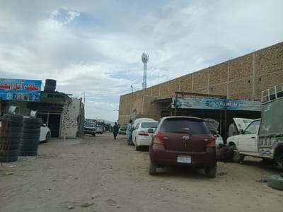The image depicts a street scene in Bostan, Pakistan. The street is unpaved and dusty, lined with small shops or workshops. In the foreground, a dark red Toyota Yaris is parked, with the license plate "BBD 519" visible. Several other cars are parked or being worked on, with hoods open. People are present, some appearing to be mechanics working on the vehicles. A tower stands in the background, constructed of metal bars and a gray/blue box with several antennas at the top of it. A large brick wall is seen running along the side of the street. On the left, a tire shop with stacked tires is visible, with a sign in Urdu that reads "Malik Oil Agency". Another sign in Urdu advertises "Old Spare Parts" and "Ziarat Road Kuchlak".
