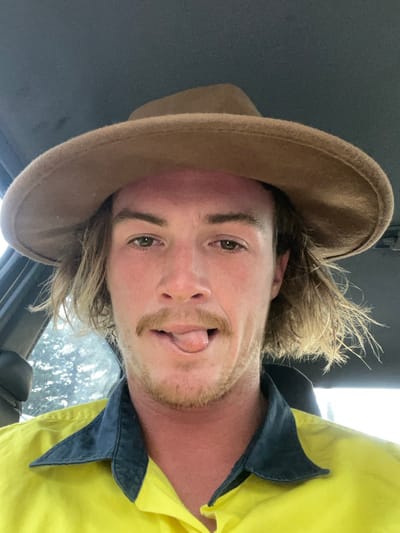 A man wearing a tan cowboy hat and yellow work shirt with dark blue collar poses with his tongue sticking out. He has blonde hair and a reddish complexion. The photograph appears to be taken from inside a vehicle, as the interior is visible. The image is rotated, presenting the subject at an angle.
