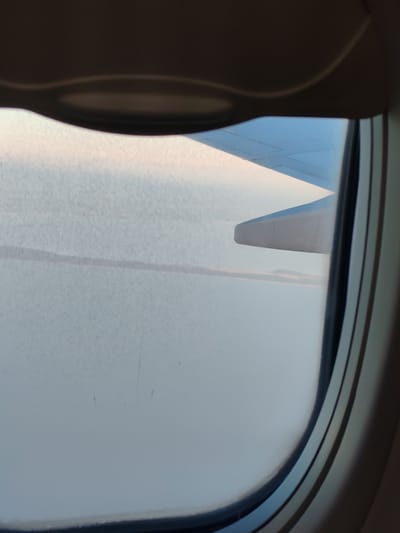 The image is taken from an airplane window, looking out at a cloudy sky and a wing. The view suggests the airplane is in flight, possibly over land given the hint of a distant horizon. There is no visible indication of the city of Khrabrovo, Russia, or any specific urban elements within the frame. The scene is predominantly sky and airplane wing, with a somewhat hazy or overcast atmosphere. The window frame of the aircraft is also visible.
