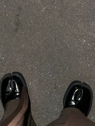 The image shows a downward shot of a person wearing shiny black Tabi shoes with dark brown pants on an asphalt surface. The shoes have a distinct split-toe design. The asphalt has a dark gray, gritty texture. The lighting is dim, casting shadows on the pants and shoes, emphasizing their glossy surface.