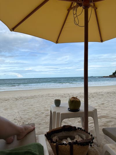 This media file depicts a relaxing scene on a sandy beach in Ao Cho, Thailand. The perspective is from someone sitting under a large, yellow beach umbrella, with the camera angled upwards and to the side. The foreground is dominated by the underside of the yellow umbrella, supported by a wooden pole.
To the right of the umbrella, the view opens up to the beach and the ocean. A white plastic table is situated on the sand, with a whole coconut on top, suggesting a refreshing drink. Next to the coconut, there's a small green cup. A woven straw bag with decorative trim rests on the table. Further to the right, a person's feet are visible, resting on what appears to be a beach chair or lounger, with a green towel draped nearby. The sand is a light color with visible ripple patterns.
The ocean stretches out in the background, appearing calm and serene with gentle waves. The sky is a bright blue with scattered white clouds, indicating a pleasant, possibly sunny day, likely in the late morning or early afternoon given the brightness. The overall mood conveyed is one of leisure and relaxation. There is no visible text in the image.