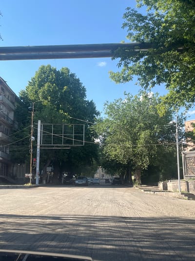 The image depicts an outdoor scene under a clear blue sky, likely during the daytime with bright sunlight casting shadows. The perspective is slightly tilted, creating a dynamic feel to the composition.

Dominating the left side of the frame are two large, thick pipes, possibly part of an industrial infrastructure or utility lines. Their weathered, dark surface contrasts with the vibrant green foliage of large trees that hang over and surround them. These trees, with their dense canopy and lush leaves, suggest a season of growth, likely summer.

To the right, a textured, light-colored asphalt road stretches into the distance. The road exhibits a rough, gravelly surface with visible tire tracks and striations, indicating it might be a less-traveled or rural road. Long, parallel shadows stretch across the road, cast by unseen objects, reinforcing the impression of strong sunlight.

Further down the road, a traffic signal with a red light illuminated is visible, along with a metal grid structure, possibly a billboard frame or a support for overhead wires. Several cars are parked along the roadside in the middle ground, and buildings with residential architecture are partially visible in the background. The overall impression is a street scene in a town or city, with a blend of natural elements and man-made structures.

The image appears to be taken from a slightly elevated or angled viewpoint, looking down the road and towards the trees. There is no clear activity or event taking place, suggesting a snapshot of a typical moment in this location. The setting hints at a residential or semi-urban area in Gladzor, Armenia, given the context provided. The absence of people makes it difficult to infer any specific emotions, but the bright sunlight and green foliage contribute to a generally pleasant, outdoor atmosphere.