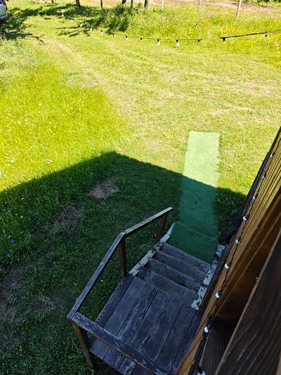 The image depicts an outdoor scene with a wooden staircase leading down to a grassy area. The staircase is made of dark, weathered wood and has a wooden handrail. The grass is green and appears to be a lawn, with some patches of drier grass or straw. A shadow is cast on the grass from an unseen object, possibly a building or structure. In the background, there is a string of lights with white bulbs stretched across the scene. The overall environment appears to be a backyard or garden area. The location is specified as Ukeshi, Georgia.