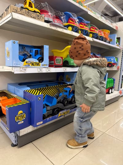 A young child wearing a brown checkered hat, a green jacket with a fur-lined hood, and blue jeans stands in front of a shelf in what appears to be a retail store, likely in the city of Korobovo, Russia, based on the presence of Russian language text on the product packaging. The child is leaning over and reaching into a box containing blue toy construction vehicles. The shelf is stocked with various other toys, including plastic vehicles in nets and boxed sets with the words "СТРОИТЕЛЬНАЯ ТЕХНИКА ACTIVE" (Construction Equipment Active) prominently displayed in Russian. Price tags are visible on the shelf edges, indicating the cost of the items. The floor is light-colored tile. The overall scene suggests a consumer environment, specifically the toy section of a store.