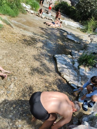 This image depicts a group of people bathing in a shallow, rocky stream with a gentle current. The stream appears to be a natural watercourse flowing over light-colored, smooth stones and gravel. Lush green vegetation lines the banks of the stream, and some of the rocks are covered in moss. In the background, there are indications of a natural, possibly mountainous, landscape.

In the foreground, a man wearing black swim trunks is bending over, apparently washing his hands or face in the water. To his right, a woman in a blue shirt and dark shorts is also interacting with the water, holding a plastic bottle. Next to her are a pair of black and white striped slides. Further upstream, several other individuals are visible, some standing in the water and others lounging on the rocks. One person is wearing red swim trunks, another is in a bikini, and a couple is lying down on the rocks near the water's edge. The overall impression is of a relaxed, outdoor recreational activity in a natural setting.

The location is identified as the Pou Clar natural pools in Ontinyent, Spain. These pools are known for their clear water, sculpted rock formations, and surrounding natural beauty, making them a popular spot for swimming and picnicking during warmer months.