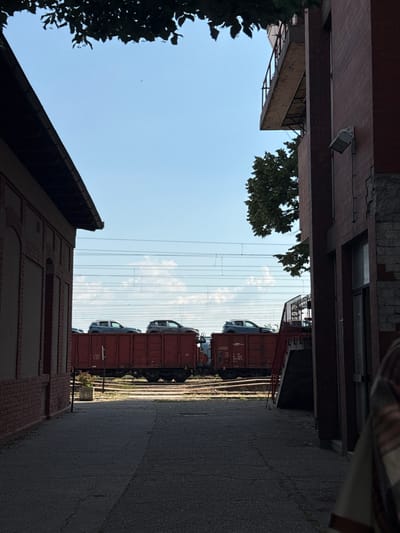 An alleyway in Ruma Municipality, Serbia, framed by brick buildings and a patch of sky with power lines visible. A train loaded with cars is on the tracks at the end of the alleyway.