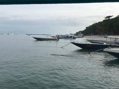 The image depicts a coastal scene in Toyapakeh, Indonesia, under an overcast sky. The foreground is dominated by calm, rippled dark green water. In the immediate foreground, the bow of a partially visible white boat is present on the bottom right. Adjacent to it, a dark blue motorboat, labeled "ARI CRUISER" on its starboard side, is moored, with dark ropes extending from its bow into the water. Behind it, closer to the sandy shoreline on the right, another white motorboat is partially visible.
Further into the water, towards the left of the frame, a dark grey speed boat with a white canopy, labeled "NUSA CENINGAN" on its side, is positioned, facing right. It has an outboard motor visible at its stern. In the far distance on the left horizon, several large vessels, including a ferry, are present. A long, dark pier extends into the water from the distant left.
The right side of the image features a light-colored sandy beach line, backed by a dense treeline of tropical vegetation. Several smaller boats are pulled up onto the beach or are very close to the shore. A tall, white, pole-like structure, possibly a navigation beacon or small lighthouse, stands near the treeline in the mid-background. The sky is uniformly grey with cloud cover. No individuals are clearly discernible or performing specific actions.