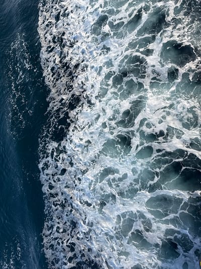 The image displays an overhead perspective of a body of water near Palaiopoli, Greece, specifically depicting a vessel's wake. The scene is bisected by a distinct vertical boundary. On the left, approximately one-third of the frame shows dark blue, relatively undisturbed water with subtle surface variations. The central and right two-thirds of the frame are dominated by a turbulent mass of white foam and aerated, light blue-green water. This foamy area exhibits complex, irregular patterns of bubbles and swirling currents, consistent with the immediate aftermath of a moving object, likely a boat or ship. No landmasses, vessels, people, or other distinct objects are visible within the frame. The lighting suggests an overhead source, illuminating the white foam prominently against the darker water.