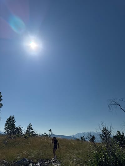The image depicts a landscape scene under a bright, clear blue sky. In the upper left quadrant, the sun is visible, creating a lens flare effect with streaks of light extending downwards and to the left. The environment appears to be a hilly or mountainous terrain with dry, golden-brown grass covering the ground. In the mid-ground, a woman is walking through the tall grass. She is wearing a dark-colored athletic outfit consisting of a crop top and shorts, with white socks and dark shoes. Her body is positioned facing towards the left side of the image, and her head is tilted downwards as if looking at the ground. To the left of the woman, several trees with green foliage are scattered across the landscape. In the background, a range of mountains is visible, appearing hazy due to the distance. The overall scene suggests an outdoor activity, possibly hiking or walking, in a natural, undeveloped area. The location is identified as Bukovac, Montenegro.
