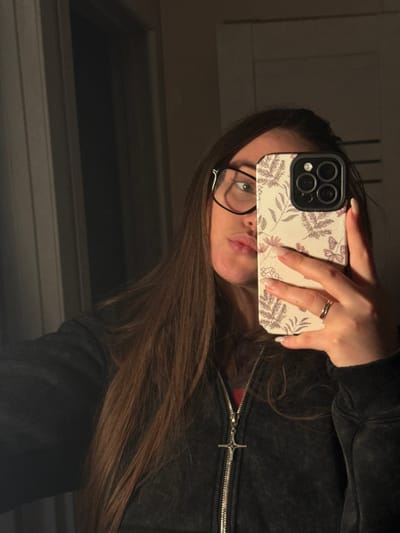 An image taken in an interior setting in Prikubanskiy, Russia, shows a female individual from the chest up, capturing a mirror selfie. The subject possesses long, straight brown hair falling over her shoulders and wears black-framed glasses. Her left eye is visible, directed towards the camera, while her lips are pursed in a pout. She is attired in a dark, possibly charcoal or black, zipped jacket, revealing a hint of a red garment underneath. The jacket's zipper features a silver-colored, stylized cross pendant. The individual holds a smartphone in her right hand, obscuring the lower right portion of her face. The phone is equipped with a light-colored, vertically ribbed case adorned with a pattern of dark red leaves and flowers. The phone's triple-lens camera array is visible. A thin, double-banded metallic ring is present on the ring finger of her right hand, and her fingernails exhibit a light-colored polish with white tips. The background is dimly lit, revealing a plain wall or door surface to the right and a dark vertical element, possibly a door frame or mirror edge, to the left.