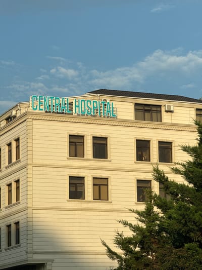 An outdoor, low-angle, vertical shot captures the upper facade of a light-yellow, multi-story building with a dark roof. The building is illuminated by sunlight, casting shadows. A bright teal neon sign across the top of the building reads "CENTRAL HOSPITAL". The sky above is blue with scattered white clouds. To the right, the upper branches of a lush green conifer tree obscure part of the building. Windows on the building are dark, suggesting they are reflecting the sky or are darkened from within. An outdoor air conditioning unit is visible on the top story of the building. The architecture appears modern, with clean lines and decorative trim. The scene is static and devoid of any people or activity. The location is identified as a street in Baku, Azerbaijan.