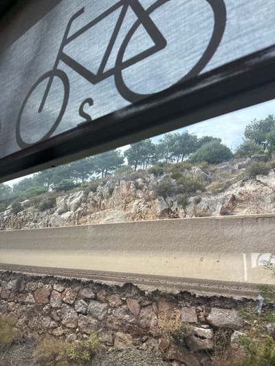 The image shows a view through a screen with a bicycle graphic overlay. Beyond the screen, a rocky cliff face is visible, covered in sparse vegetation and trees. To the right of the cliff, a paved surface, possibly a road or pathway, runs alongside. The ground next to the paved surface is rocky and covered with dry grass and some small plants. The sky is cloudy. The scene appears to be outdoors, likely in a natural or semi-natural setting. The time of day is not explicitly indicated, but the lighting suggests it is daytime. The location is identified as Salses-le-Château, France. There is no visible text in the image, other than the bicycle graphic on the screen.
