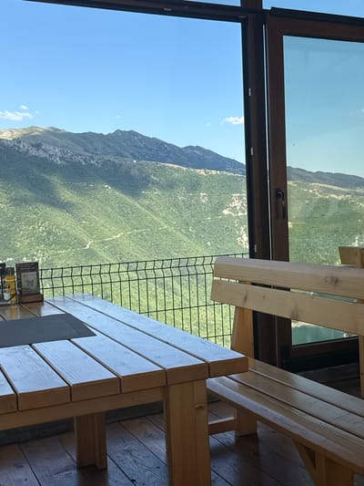 The image depicts a view from an elevated structure, possibly a restaurant or viewing platform, overlooking a lush green mountainside. The scene is dominated by natural elements, with the rugged terrain and vibrant vegetation stretching out below. In the foreground, elements of the man-made structure are visible, including wooden tables and benches, suggesting a place designed for visitors to enjoy the scenery. A dark metal fence separates the seating area from the expansive view, adding a layer of safety while not obstructing the panorama.
The sky above is a clear, bright blue, indicating fair weather and likely daytime. The lighting suggests it could be late morning or early afternoon, with the sun casting shadows on the mountainside, highlighting its contours and texture. The overall impression is one of tranquility and natural beauty, a serene escape in a mountainous region.
A small sign with a QR code is visible on one of the tables, likely for menu access or information. Some of the text on the sign is legible, reading "VREMENSKI USLOVI NAGRADJUJE," which translates from Croatian/Serbian to "Weather conditions reward." This hints at the location being a place where enjoying the weather is part of the experience. The setting evokes a sense of peace and awe, inviting contemplation of the vast natural landscape.