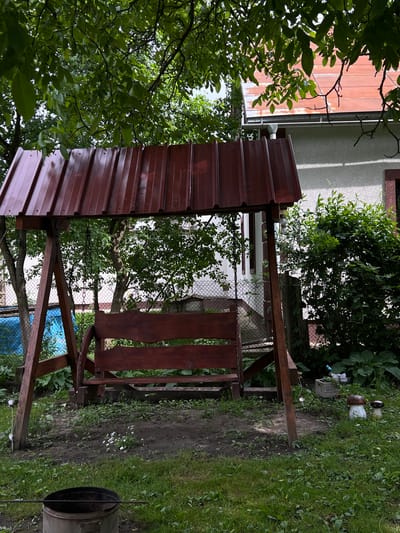 The image depicts a backyard scene in Mshanets, Ukraine, featuring a wooden garden swing with a reddish-brown corrugated metal roof. The swing structure is made of dark-stained wood, and the bench is suspended by chains.
In the foreground, the ground is a mix of sparse grass and exposed soil patches, with small white flowers visible in one dirt area. A dark, cylindrical metal object, possibly a bucket or fire pit, with a metal rod resting across its top, is positioned in the lower-left corner. Towards the right foreground, two mushroom-shaped garden ornaments and a small planter box containing a butterfly decoration are visible on the grass.
The midground is dominated by the swing set. Behind the swing on the left, a blue tarp or cover is partially visible through green foliage and a chain-link fence. Trees and dense green bushes frame the scene, with a chain-link fence running horizontally in the background, partially obscured by vegetation.
In the background, a light-colored building, likely a house, is visible. It features a reddish-orange tiled roof with a gutter and downspout system. A window with a dark frame is also discernible on the house wall. Overhead, lush green leaves from mature trees create a dense canopy, partially obscuring the sky. No people are present in the scene.