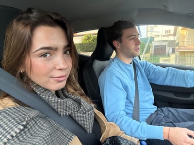 The scene depicts two young adults occupying the front seats of a moving vehicle within a residential area of Mafra, Portugal. A female passenger, positioned in the foreground-left, has long brown hair and wears a beige jacket over a dark garment, accessorized with a gray and beige houndstooth scarf. She is secured by a dark seatbelt across her chest and lap, and directly faces the viewer with a slight smile. To her right, a male driver is observed in profile, focused forward on the road. He wears a light blue long-sleeved sweatshirt and dark trousers, with a red string bracelet visible on his left wrist. He is also secured by a seatbelt. The car's interior features dark upholstery. Through the vehicle's windows, the exterior environment reveals multi-story, light-colored buildings with tiled roofs, interspersed with green foliage, consistent with a daytime setting.
