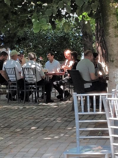The image shows a group of people sitting at tables outdoors, likely at a restaurant or cafe. The scene is set with trees and foliage in the background, suggesting a pleasant, possibly warm day. The people appear to be engaged in conversation and enjoying a meal or drinks. There are several tables with chairs, and some of the tables have items on them that could be food or drinks, though they are not clearly identifiable. The people are dressed casually, and their expressions are not distinct enough to ascertain specific emotions. A white structure, possibly a fence or railing, is visible on the right side of the image. The lighting suggests it is daytime, with sunlight casting shadows. No visible text is present in the image. The overall impression is a relaxed social gathering in an outdoor setting.
