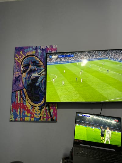 The scene depicts an interior space in El Hueco, Argentina, featuring a wall-mounted television, a laptop, and a large piece of artwork. A flat-screen television on the right displays a live football match, with a scoreboard indicating "83:56 RMA 3 0 MCI" and "crypto.com" sponsor banners visible on the pitch. Below and to the right of the television, an open laptop, possibly a gaming model with a backlit keyboard, shows a different football match, with a scoreboard displaying "10:29 PSG 1 0 CAE" and an "ESPN" logo. A player wearing a white jersey with the number "10" is prominently visible on the laptop screen. To the left of the television, a large, rectangular canvas print depicting a colorful, graffiti-style portrait of The Notorious B.I.G. (Biggie Smalls) is affixed to the wall. The artwork portrays the subject wearing a crown, a dollar-sign pendant chain, and smoking, with fragmented text elements like "NEVER LET THEM YOUR" and "NOTORIOUS" integrated into the design. The wall behind the screens and artwork is painted a uniform grey. A black cable extends downward from the rear of the television. An indistinct, dark object is partially visible at the bottom left of the frame.