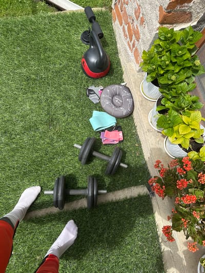 The scene is outdoors, possibly a backyard or patio, in Rogozinovo, Bulgaria. The ground is covered in artificial turf. In the upper portion of the frame, there are several potted plants: two with green leafy foliage and one with small orange flowers.
Arranged on the turf are various fitness-related items: an ab roller, a weighted plate (marked “Bakkin 4 KG”), exercise resistance bands, exercise gloves, and two dumbbells.
In the lower right corner, a pair of feet wearing white socks and the lower portion of red and black athletic pants are visible, suggesting a person standing in the scene.